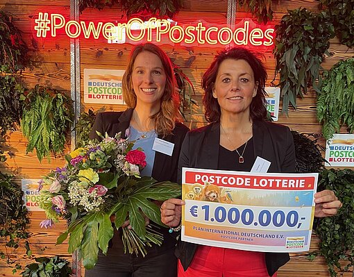 Claudia Meyerhöfer und Petra Berner stehen nebeneinander, eine hält einen Blumenstrauß in der Hand, die andere einen Scheck in Höhe von 1 Million Euro