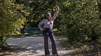 Eine junge Frau steht auf einem Schotterweg, hat eine Hand in die Hüfte gestemmt und hebt die andere Hand als Faust über ihren Kopf
