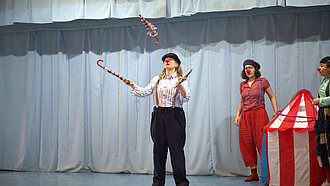 Drei Clowns stehen auf einer Bühne. Eine von ihnen jongliert mit drei Regenschirmen.