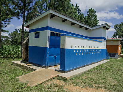 Ein kleines, schlichtes Gebäude mit blauer und weißer Bemalung – eine Schultoilette – steht auf einer grünen Wiese, umgeben von Bäumen und Feldern.