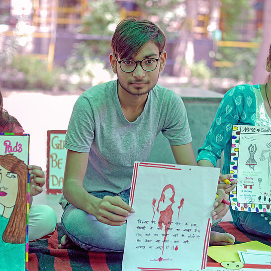 Kinder sitzen in Indien unter einer Zeltplane auf Decken und zeigen Plakate über Menstruationshygiene vor