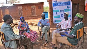 Fünf Personen, darunter Männer und Frauen, nehmen an einer Besprechung im Freien teil, im Hintergrund ist ein Plakat der Organisation "Plan International" zu sehen