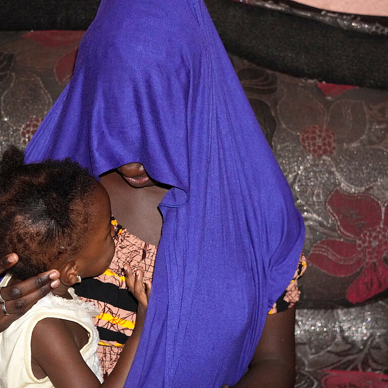 Fatoumata trägt ein Tuch über dem Kopf, sodass man ihr Gesicht nicht erkennen kann. Sie hält ihre kleine Tochter im Arm