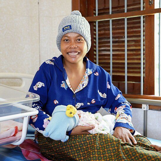Eine frisch entbundene Mutter sitzt im Gesundheitszentrum, hält ihr Baby im Arm und lächelt glücklich in die Kamera.