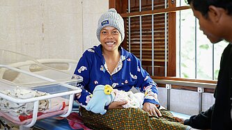 Eine frisch entbundene Mutter sitzt im Gesundheitszentrum, hält ihr Baby im Arm und lächelt glücklich in die Kamera.