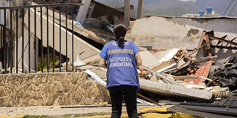 Eine Frau steht vor einem zerstörten Haus. Sie steht mit dem Rücken zur Kamera und trägt eine blaue Weste mit dem Logo von Plan International. 