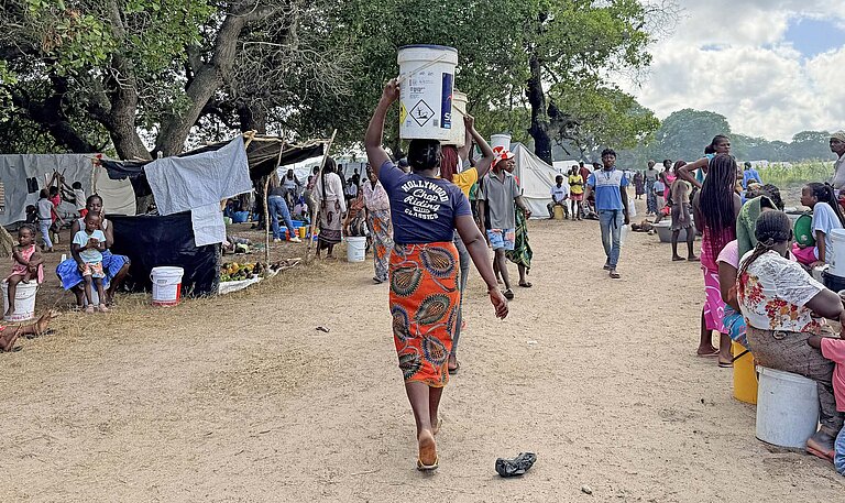 Eine Frau balanciert Güter auf dem Kopf und läuft durch ein Notcamp aus Zelten
