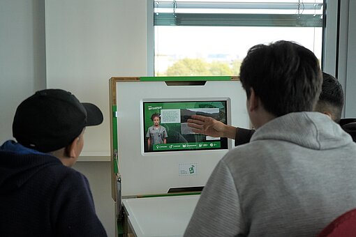 Drei Jungen sitzen vor einer Box mit Bildschirm, einer zeigt auf das, was auf dem Screen zu sehen ist.