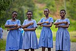 Vier ghanaische Schulmädchen in blaukarierten Schuluniformen stehen nebeneinander im Freien und lächeln in die Kamera. Sie halten ihre Unterarme vor ihrem Körper, sodass sie ein Gleichzeichen bilden.. Die Umgebung ist grün und ländlich.
