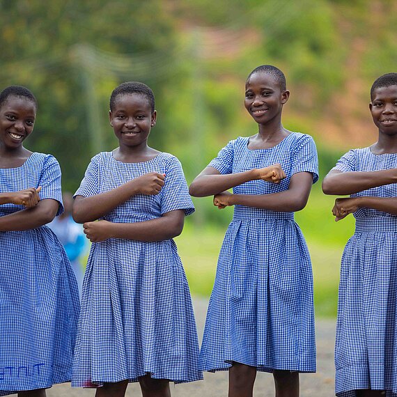 Vier ghanaische Schulmädchen in blaukarierten Schuluniformen stehen nebeneinander im Freien und lächeln in die Kamera. Sie halten ihre Unterarme vor ihrem Körper, sodass sie ein Gleichzeichen bilden.. Die Umgebung ist grün und ländlich.