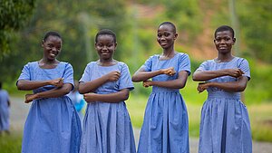 Vier ghanaische Schulmädchen in blaukarierten Schuluniformen stehen nebeneinander im Freien und lächeln in die Kamera. Sie halten ihre Unterarme vor ihrem Körper, sodass sie ein Gleichzeichen bilden.. Die Umgebung ist grün und ländlich.