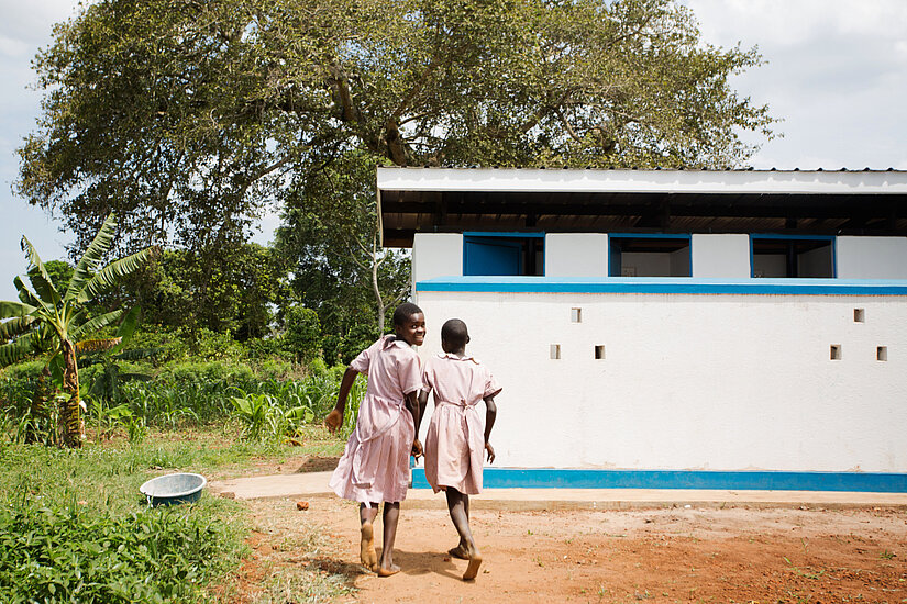 wei Schulmädchen in Uniform laufen lachend zu einer modernen Schultoilette in ländlicher Umgebung in Uganda – Zugang zu sanitären Einrichtungen fördert Schulbesuch.