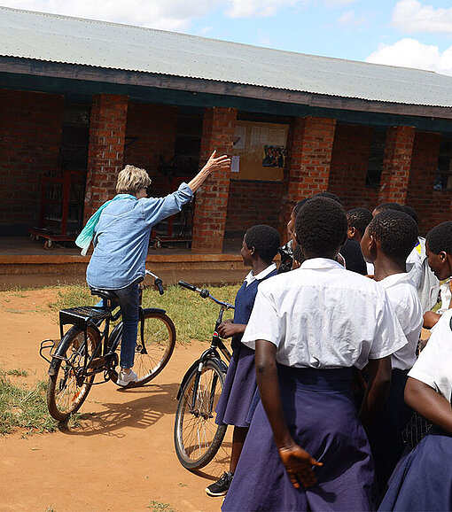 Besucherin fährt ein Schulrad auf dem Schulhof in Malawi, Kinder schauen zu.