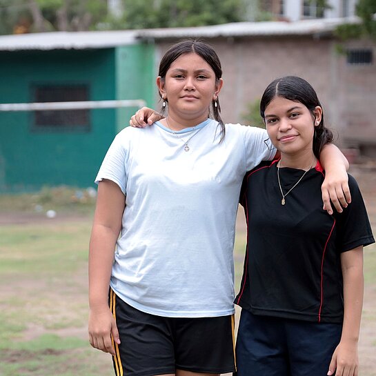 Virginia und Saidia stehen Arm in Arm draußen auf dem Fußballplatz ihrer Gemeinde