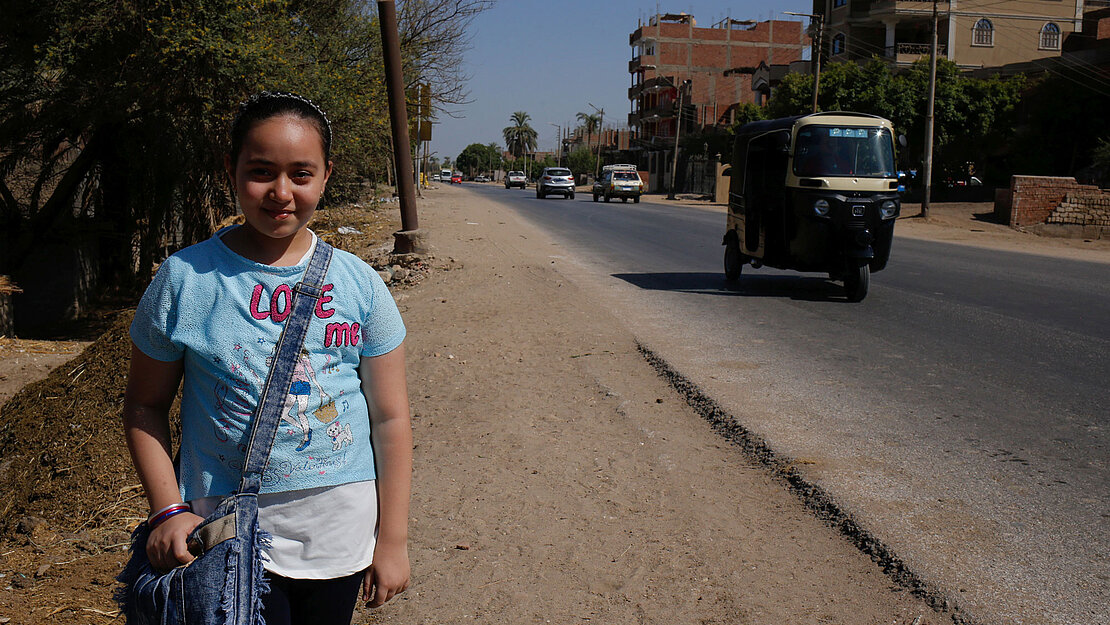 Ein Mädchen mit einem blauen T-Shirt steht am Straßenrand und lächelt in die Kamera.