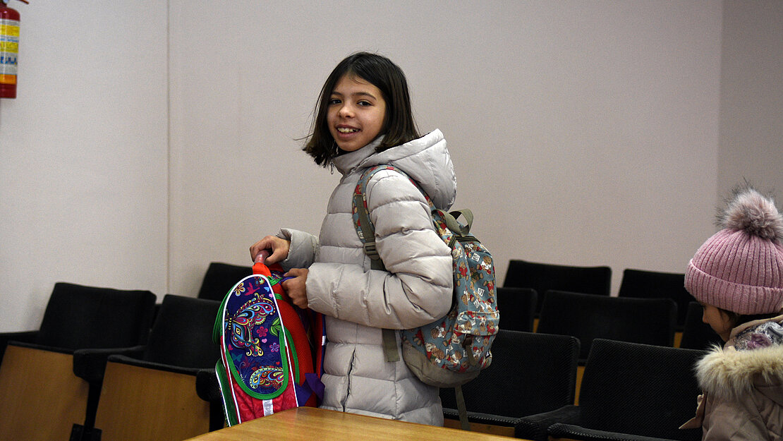 Ein Mädchen hat einen Schulranzen in der Hand und lacht in die Kamera.