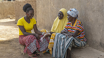 Drei Frauen sitzen im Schatten eines Hauses, die jüngste hat eine Damenbinde in der Hand.
