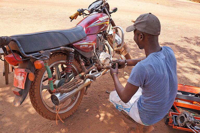 Ein junger Mann kniet vor einem Motorrad, dass er repariert.