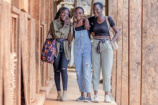 Drei junge Frauen vom Upcyclers Creative Hub gehen gemeinsam auf einem Weg mit Holzwänden in Mathare, Kenia. Die Initiative stärkt Frauen und kämpft gegen Periodenarmut.