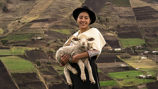 Ein Mädchen steht mit einem Schaf auf dem Arm vor Feldern in Ecuador.