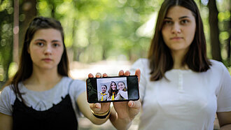 Zwei junge Frauen stehen nebeneinander und zeigen ein Handy in die Kamera, auf dem ein Bild mit drei Menschen zu sehen ist