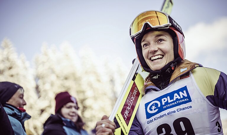 Eine Skispringerin in Wettkampfkleidung trägt ihre Ski auf der rechten Schulter. Sie lacht.