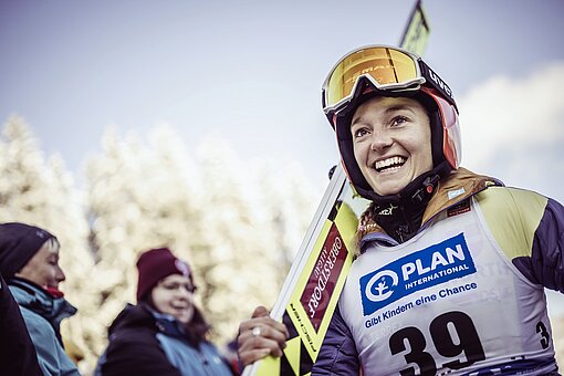Eine Skispringerin in Wettkampfkleidung trägt ihre Ski auf der rechten Schulter. Sie lacht.