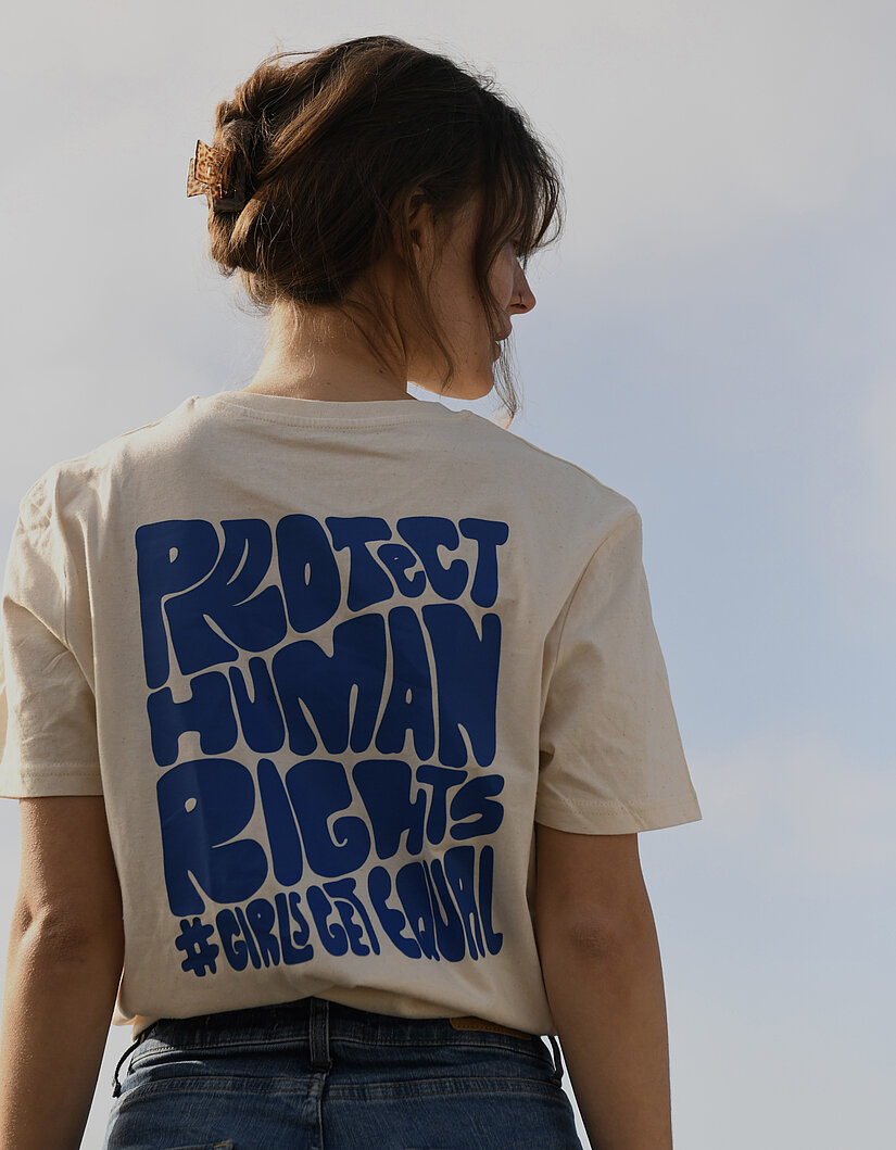 Hannah ist von hinten fotografiert. Sie trägt ein blau bedrucktes T-Shirt, mit der Aufschrift "Protect Human Rights #GirlsGetEqual"
