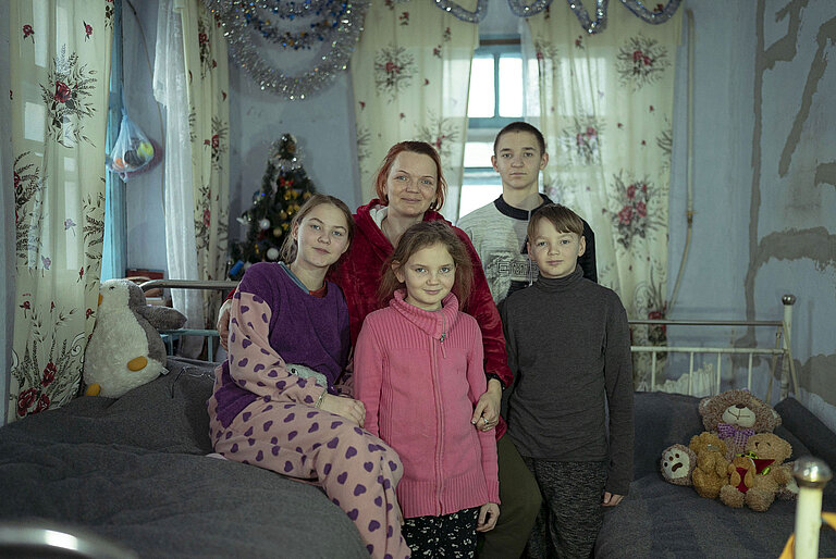 Eine Frau steht mit ihren vier Kindern in einem Zimmer zwischen Betten. 
