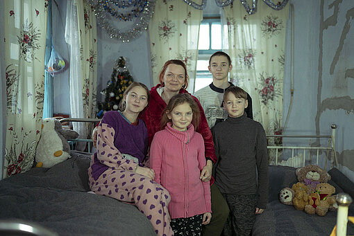 Eine Frau steht mit ihren vier Kindern in einem Zimmer zwischen Betten. 