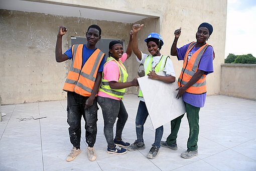 Drei Frauen und ein Mann stehen auf einer Baustelle und strecken die Arme in die Luft