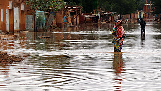 Eine junge Frau in buntem Gewand läuft durch eine überflutete Straße. Sie hält ihre Schuhe in der Hand.