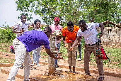 Mehrere Menschen, darunter Vertreter:innen von Plan International, testen gemeinsam einen neu gebauten Wasserbrunnen in einem ländlichen Dorf in Ghana. Sie lassen Wasser aus dem Pumpbrunnen laufen und fangen es in den Händen auf. Im Hintergrund sind weitere Personen, Bäume und einfache Hütten zu sehen.
