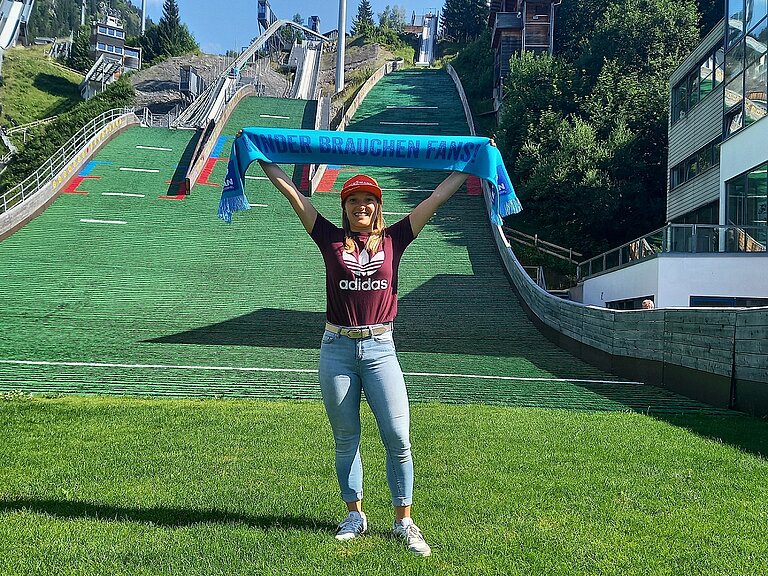 Katharina Althaus steht vor einer Sommer-Skischanze mit Rasen und hält einen „Kinder brauche Fans“-Schal hoch