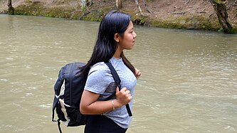 Anahi steht mit einem Rucksack am Rand eines Flusses