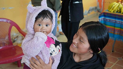Eine Frau hält ihr Baby auf dem Arm und lächelt es an. Das Baby schaut neugierig in die Kamera.