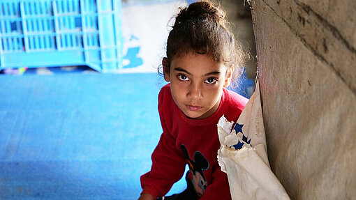 Ein Mädchen aus Syrien sitzt in einem Geflüchtetencamp auf dem Boden und spielt mit Spielzeug. Sie schaut direkt in die Kamera.