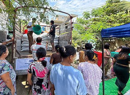 Eine Gruppe erwachsener Frauen und Männer steht in einer kleinen Menschentraube vor einem Lastwagen. Zwei Männer auf dem Wagen verteilen große Säcke und Plastikkörbe mit Hilfsgütern an die Wartenden. Die Erwachsenen tragen bunte, Alltagskleidung, viele haben ihre Haare hochgesteckt. 