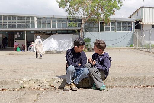 Zwei Kinder sitzen nebeneinander auf einer niedrigen Mauer im Innenhof einer Unterkunft und unterhalten sich.