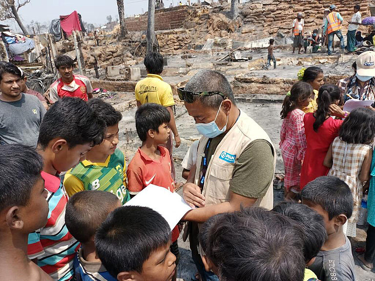 Plan-Mitarbeiter im zerstörten Camp umgeben von Kindern.