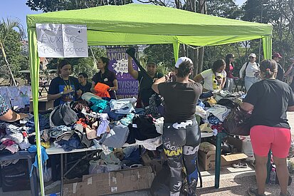 Freiwillige und Betroffene sortieren und erhalten Kleiderspenden unter einem grünen Pavillon; das Schild weist auf psychologische Hilfe hin und viele Kleidungsstücke liegen auf Tischen.