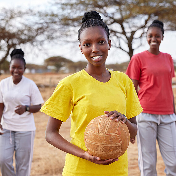 Eine junge Frau trägt ein gelbes T-Shirt und hält einen Ball, während sie im Freien steht. Im Hintergrund befinden sich zwei weitere lachende junge Frauen und Bäume in einer trockenen Landschaft.