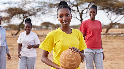 Eine junge Frau trägt ein gelbes T-Shirt und hält einen Ball, während sie im Freien steht. Im Hintergrund befinden sich zwei weitere lachende junge Frauen und Bäume in einer trockenen Landschaft.