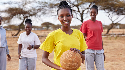 Eine junge Frau trägt ein gelbes T-Shirt und hält einen Ball, während sie im Freien steht. Im Hintergrund befinden sich zwei weitere lachende junge Frauen und Bäume in einer trockenen Landschaft.