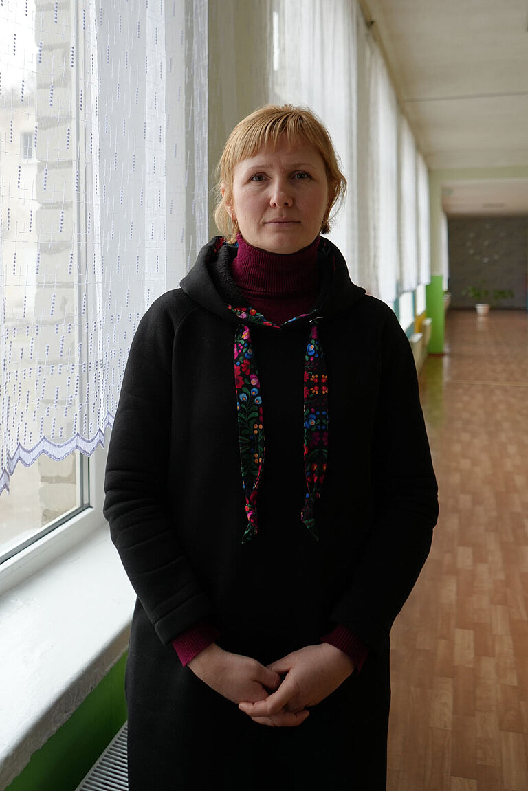 Eine Frau steht in einem hellen Schulflur. Sie schaut ernst in die Kamera.