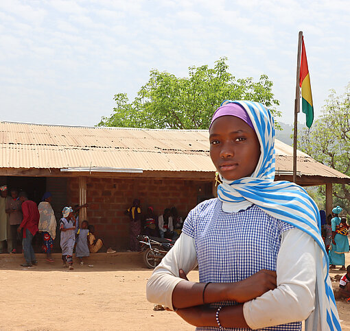 Schwarzes Mädchen mit Kopftuch mit verschränkten Armen vor baufälligem Schulgebäude