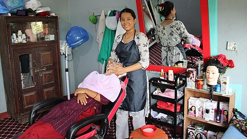 Eine junge Frau steht in einem Friseursalon und behandelt eine Kundin, die in einem Friseurstuhl sitzt. Im Hintergrund sind Friseurausrüstung und Produkte sichtbar.