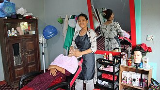 Eine junge Frau steht in einem Friseursalon und behandelt eine Kundin, die in einem Friseurstuhl sitzt. Im Hintergrund sind Friseurausrüstung und Produkte sichtbar.