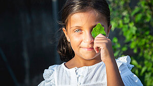 Ein Mädchen hält ein grünes Blatt vor ihr Auge und lächelt.