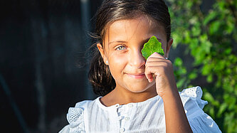 Ein Mädchen hält ein grünes Blatt vor ihr Auge und lächelt.